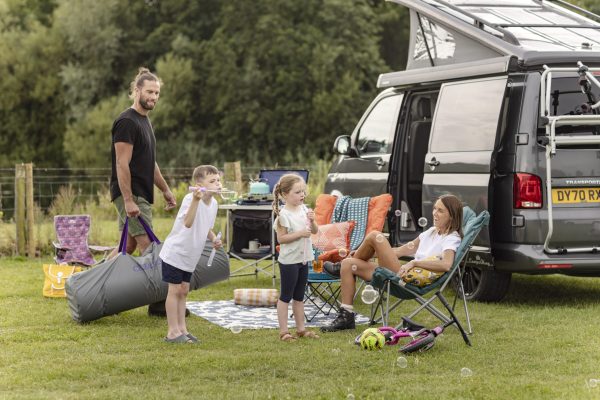 Gezin kampeert bij camperbus op camping; kinderen blazen bellen, moeder ontspant in stoel.